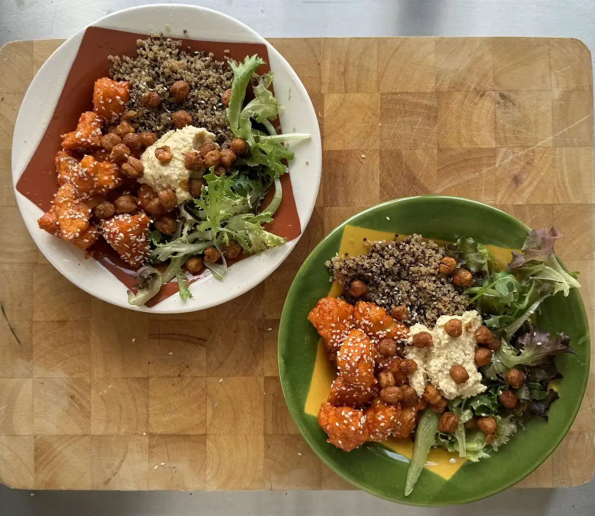 Two bowls of roasted sweet potatoes, quinoa, greens, chickpeas, and sesame seeds on a wooden board.