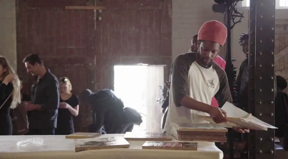 Man in a beanie browsing artwork on a table in an art gallery with people in the background.