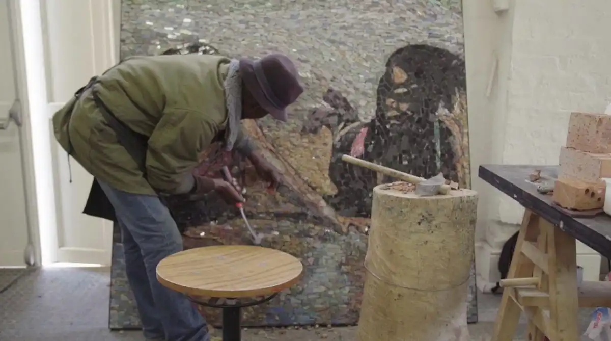 Artist working on a mosaic, wearing a hat and coat, surrounded by tools and bricks in a studio.