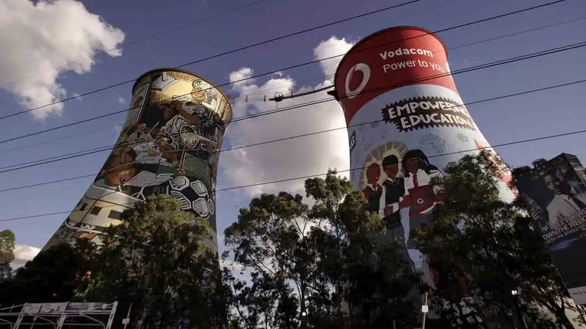 Colorful cooling towers with murals, trees, and power lines in the foreground under a partly cloudy sky.