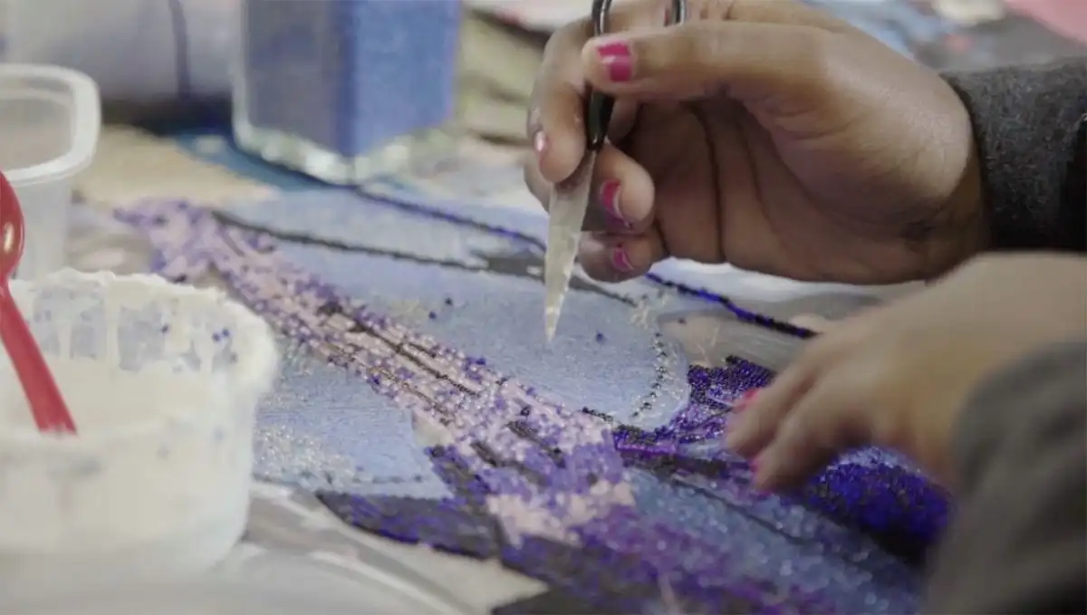 Hands using tweezers to place small beads on a detailed, colorful mosaic artwork.