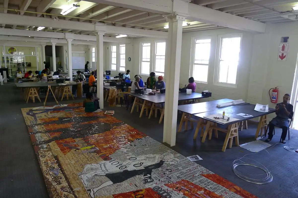 People working on a large mosaic artwork in a spacious studio with long tables and natural light.