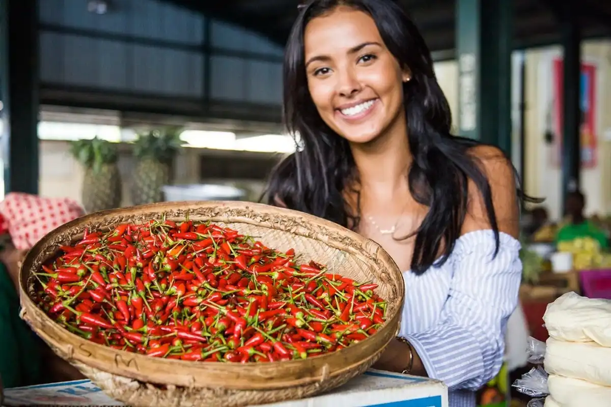 A woman smiling and holding a large basket of red chili peppers at a market.