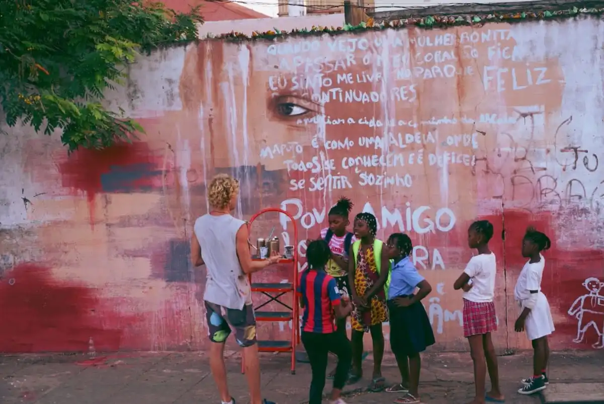 A man with curly hair paints a mural on a wall, surrounded by children watching him. The mural features text and a large eye.