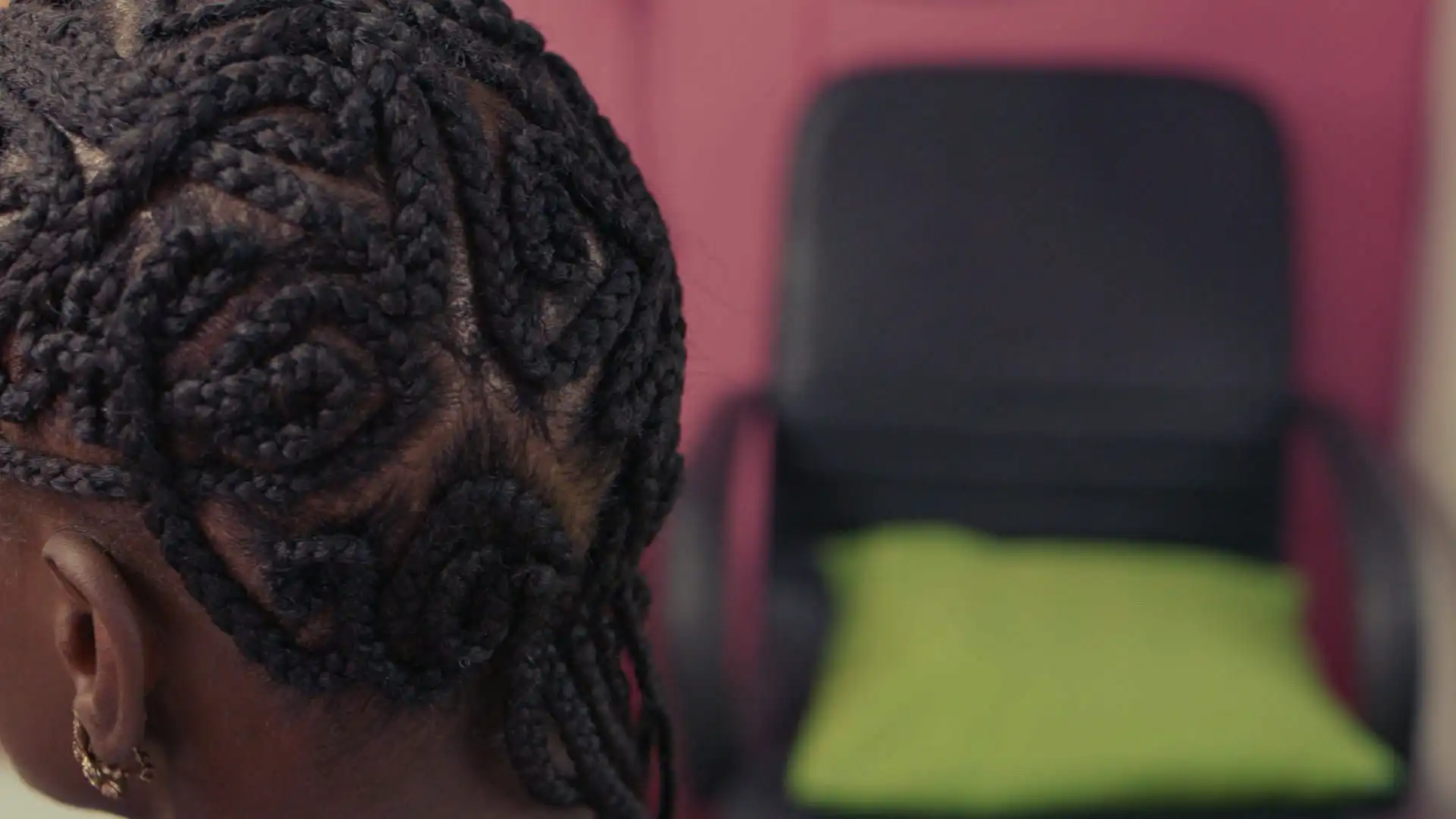 Close-up of intricate braided hairstyle with circular patterns on a person's head, with a blurred chair and green cushion in the background.