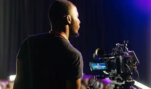 Person operating a video camera in a dimly lit setting, facing a stage with blue and purple lighting.