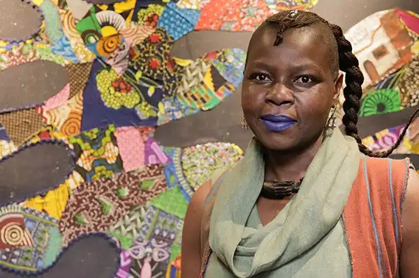 Person with braided hair and purple lipstick stands in front of a colorful, patterned textile artwork.