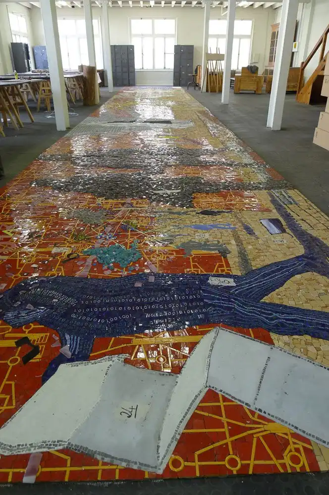 Colorful mosaic artwork on a long floor, featuring abstract designs and geometric patterns in a spacious room with white pillars.