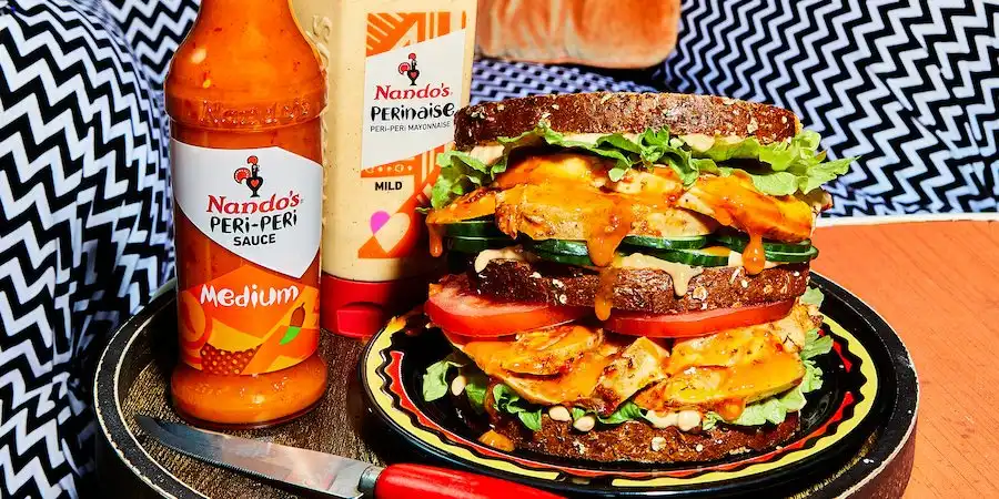 A loaded sandwich with grilled chicken, tomatoes, and lettuce, next to Nando's PERi-PERi sauce and PERinaise bottles.
