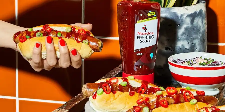 Person holding a hot dog topped with Nando's PERi-BBQ Sauce and sliced chilies. A bottle of sauce and bowl of salad are nearby.