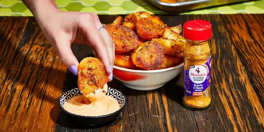 Hand dipping a seasoned potato wedge into a creamy sauce, with Nando's Garlic & Herb seasoning bottle nearby on a wooden table.