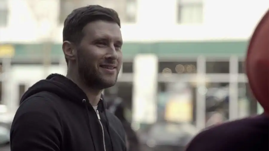 Man in a black hoodie smiling outdoors, with a blurred urban background.