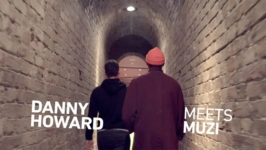Two people walking down a brick tunnel. Text reads "Danny Howard Meets Muzi."