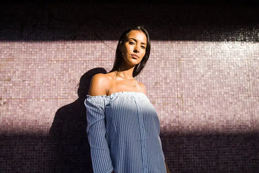 Woman in an off-shoulder striped dress stands against a textured purple wall, with sunlight casting shadows.