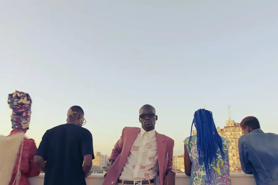 A stylish person in a pink suit and sunglasses stands confidently on a rooftop with four others, overlooking a cityscape.