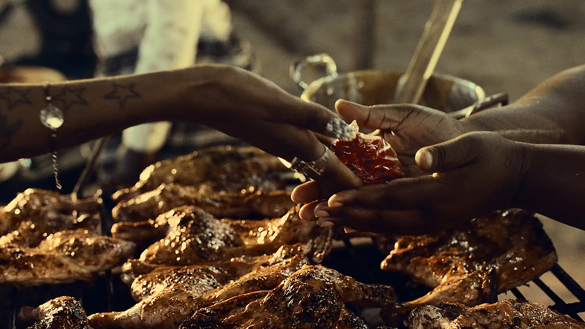 Two hands exchange a small packet over a grill with flame-grilled chicken, surrounded by cooking utensils.