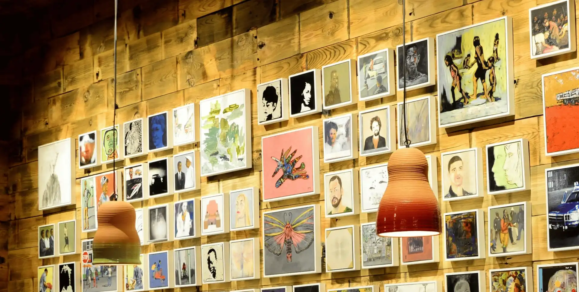 A wooden wall adorned with diverse, colorful artwork and two hanging lamps in a cozy setting.